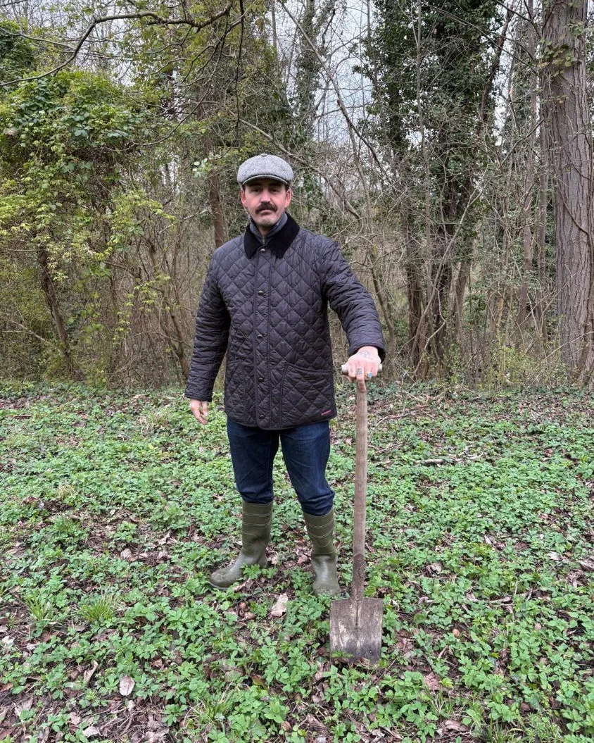 Andreas Maiorescu mit Schiebermütze und Steppjacke im Wald, auf einen Spaten gestützt