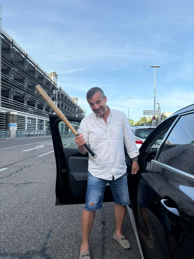 Ein Gastgeber posiert grinsend mit einem Baseballschläger an einer offenen Autotür vor einem Parkhaus