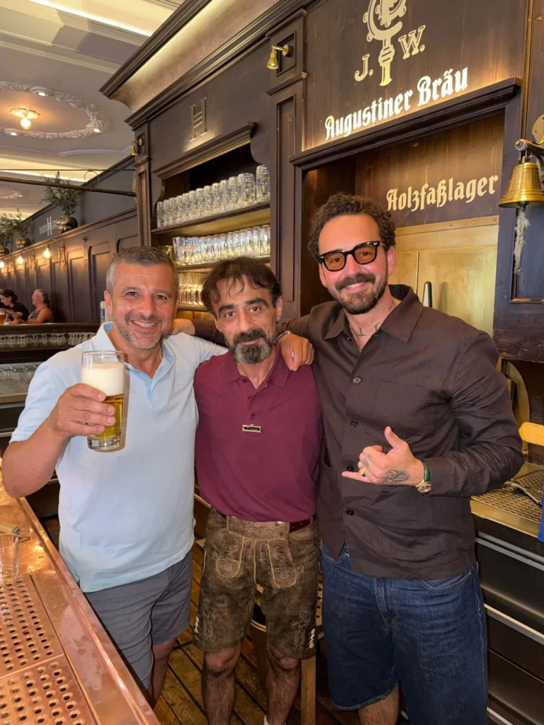 Drei Männer lachend an der Bar im Haxengrill vor dem Augustiner-Bräu-Schild, einer hebt ein Bier