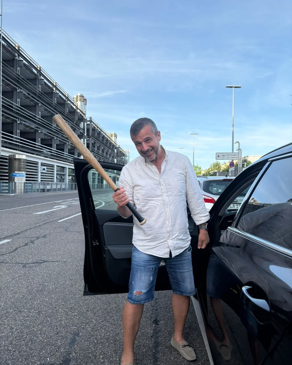 Pavlos Chatzigeorgiou grinsend mit Baseballschläger an einer offenen Autotür vor einem Parkhaus